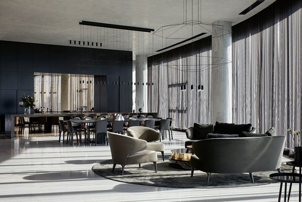 A sophisticated open-plan interior combining a formal dining area and lounge. Floor-to-ceiling sheer drapery floods the space with daylight, while dark paneled walls, pale stone or concrete surfaces, sculptural pendant lighting, and contemporary upholstered seating create a refined, monochromatic atmosphere.