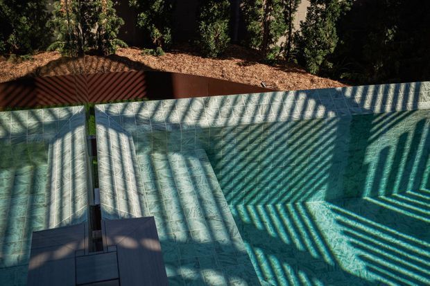 A close-up view of a swimming pool with pale blue-green tiled surfaces, largely empty of water, and strong striped shadows cast across the basin. A dark paved edge or step is visible in the foreground, with a planted wooded area and reddish-brown structure beyond the pool perimeter.