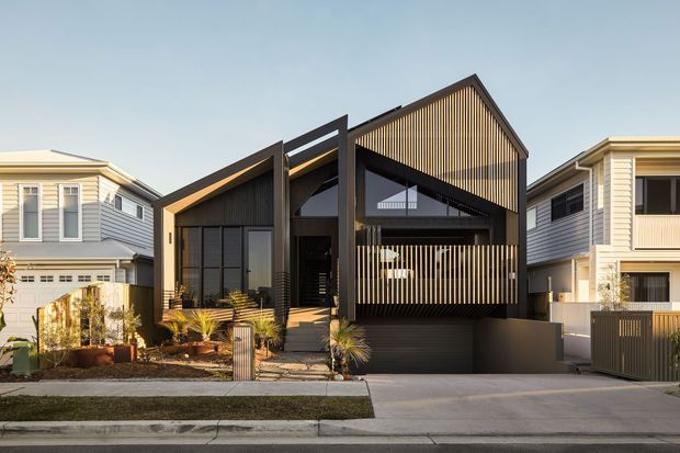 Street-facing exterior view of a contemporary multi-level house in a suburban setting. The home has a strongly geometric gabled roof form, dark vertical timber-like screening, black framed glazing, projecting structural elements, and a recessed upper balcony or terrace. Pale neighbouring homes flank the property, while a driveway and landscaped front garden occupy the foreground.