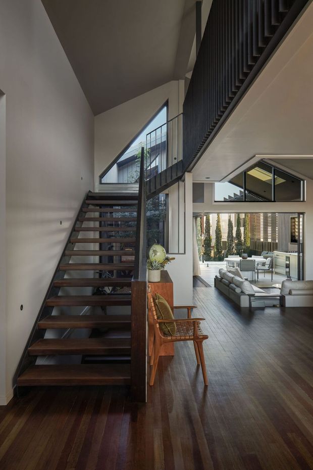 A contemporary open-plan interior viewed from a dark timber staircase. The stair has floating timber treads, a slim dark metal balustrade, and a dramatic angled ceiling. Beyond the stair is a double-height living area with dark timber flooring, pale walls, expansive glazing, a light sectional sofa, and an outdoor terrace visible through the doors. A timber console and mid-century-style chair sit beside the staircase.