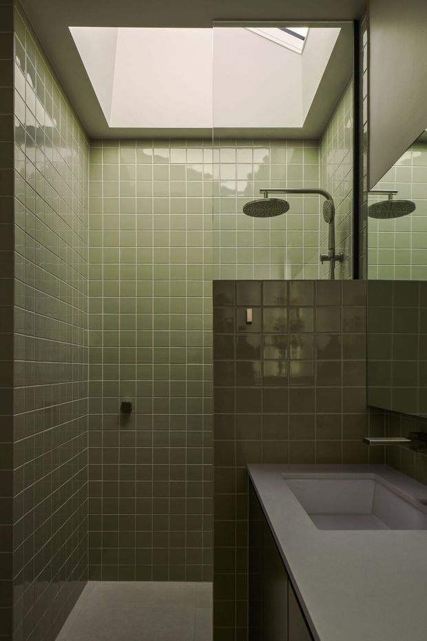 A compact, moody bathroom with a walk-in shower and built-in bathtub. Pale sage-green square tiles cover the shower walls and the partial privacy wall, while a large skylight brings in soft natural light. Dark brushed-metal shower fittings and a wall mirror contrast with the muted green tilework. The scene has a restrained contemporary feel with clean geometric lines and a partially concealed bath.