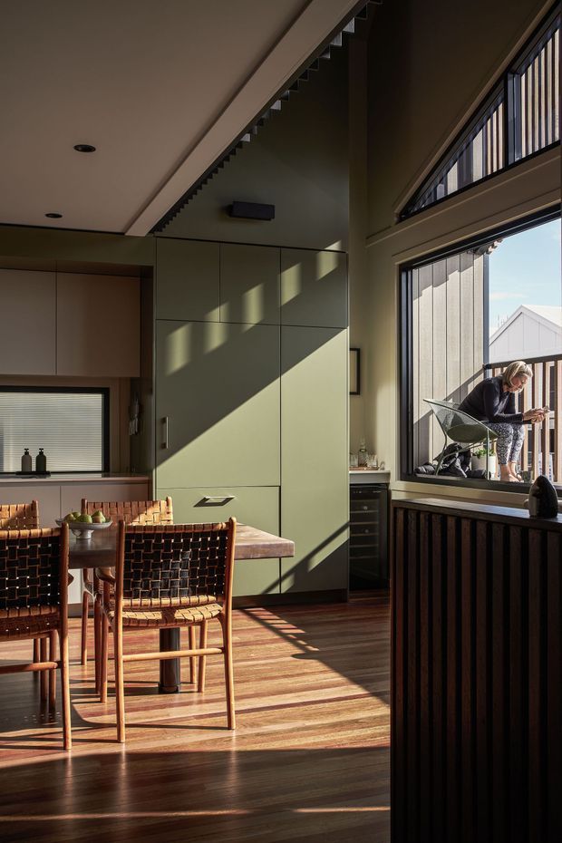 A warm, sunlit kitchen and dining area with large windows and vertical blinds. The room features dark timber flooring, muted olive-green full-height cabinetry, timber cabinetry and benchtops, and a compact dining table with woven leather chairs. Strong geometric shadows fall across the cabinetry and floor, while a person is visible outside on a balcony through the glazing.