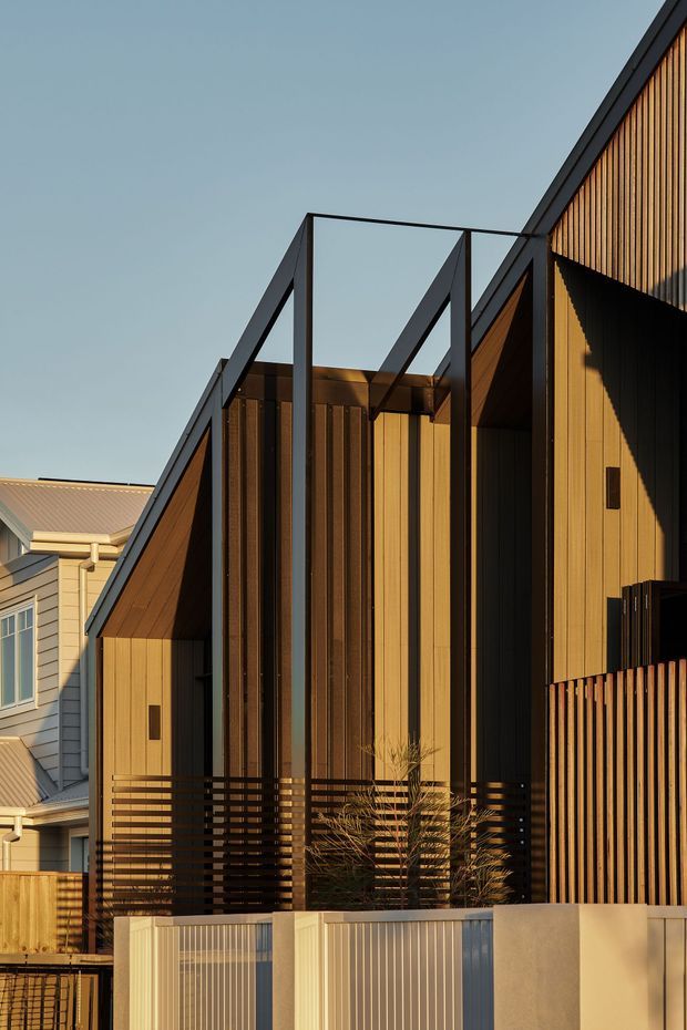 Exterior view of a contemporary multi-level residential building with a sharply articulated pitched roof and dramatic black steel framing. The facade combines warm vertical timber cladding, dark recessed openings, and a slatted timber privacy screen. A low front fence and neighbouring weatherboard housing are visible, with strong late-afternoon sunlight emphasizing the geometric roof forms.