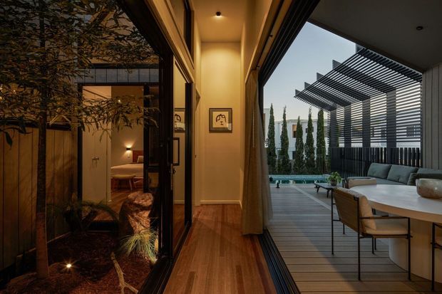 A warm, contemporary residential transition space connecting an interior hallway to a landscaped courtyard and elevated outdoor entertaining terrace. The hallway has timber flooring, warm beige walls, recessed ceiling lights, full-height glazing, and dark-framed sliding doors. Through the glazing, a bedroom-like interior is visible on the left, while the terrace on the right includes a dining table, chairs, outdoor seating, timber decking, a swimming pool, and a prominent slatted pergola and privacy screen.