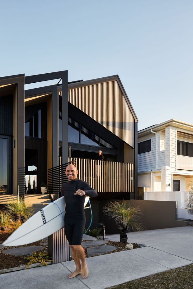 Exterior streetscape photograph of a contemporary multi-storey residence with strong geometric forms, dark framed volumes, vertical timber screening and black cladding. A lighter weatherboard house is visible beside it. The landscaped frontage includes paving, ornamental grasses and low planting. A person holding a surfboard stands in the foreground.