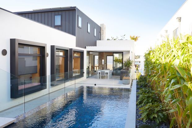Contemporary multi-level residential courtyard with a narrow swimming pool, frameless glass balustrade, white rendered walls, dark vertical cladding, and expansive glazed openings connecting to an outdoor dining and living area. Dense planting lines one side of the pool, creating a private, bright outdoor setting.