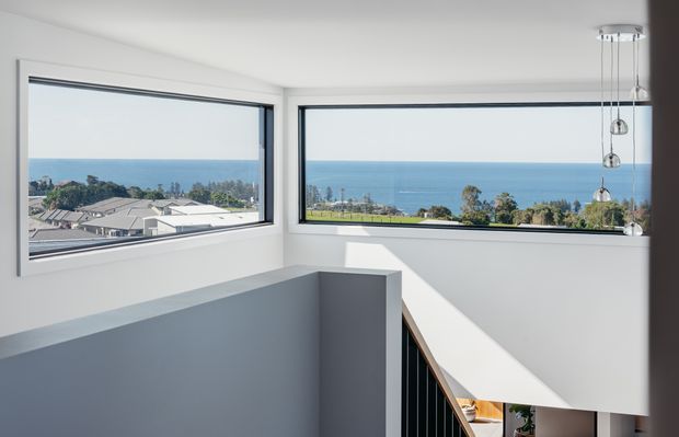 Bright, minimalist upper-level stair landing with crisp white walls, a partial-height grey stair balustrade, black vertical railing, and a contemporary cluster pendant light. Large corner windows frame an expansive ocean and coastal suburban view.