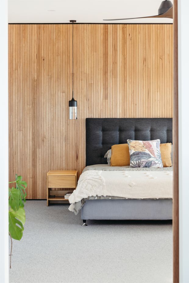 A softly lit contemporary bedroom featuring a full-height vertical timber-panelled feature wall, a charcoal upholstered bedhead, layered white bedding with mustard and botanical-patterned cushions, a small timber bedside table, pendant lighting, grey carpet, and a leafy indoor plant. A ceiling fan is partially visible overhead.