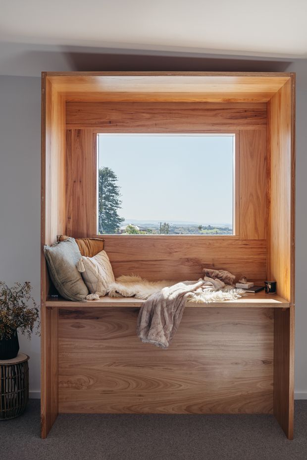 A cozy built-in timber window seat with deep wood panelling, a large picture window, layered cushions and a soft throw. The custom joinery forms a substantial framed alcove with enclosed storage below, overlooking a leafy landscape.
