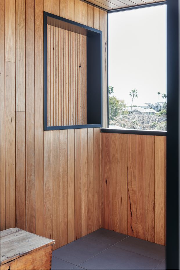 A compact timber-lined interior corner with warm vertical wood panelling on the walls and ceiling, dark charcoal tiled flooring, and a black-framed projecting window overlooking a residential landscape. A recessed timber door or cupboard is set within a dark-framed opening, with a rustic timber bench or cabinet partially visible in the foreground.