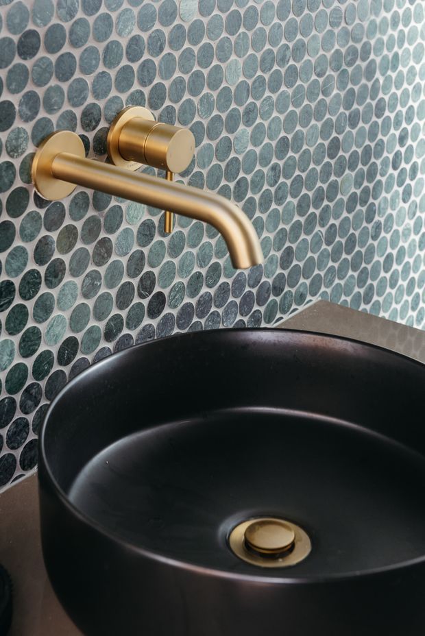 Close-up of a refined bathroom vanity featuring a matte black vessel basin, brushed brass wall-mounted tapware and a gold-toned drain set against a decorative blue-green oval mosaic tile wall. A dark stone or engineered-stone benchtop is visible around the basin.