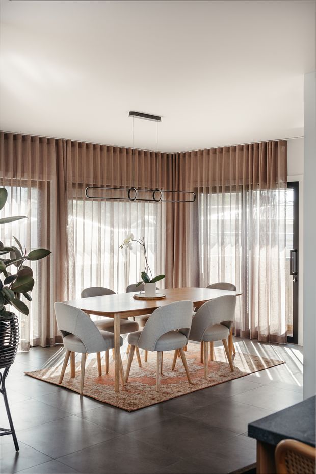Bright, refined dining area with a round light-wood dining table surrounded by upholstered dining chairs. Floor-to-ceiling corner glazing is dressed with warm taupe curtains and sheer blinds, while a slim black linear pendant hangs above the table. Large dark floor tiles, a patterned muted-rust rug, a potted plant, and strong natural light create a contemporary, warm setting.