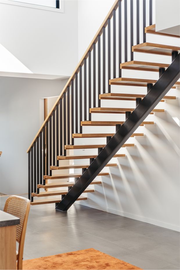 Bright contemporary interior focused on an open-riser staircase with warm timber treads, slim black vertical balusters, and a dark steel central stringer. The staircase rises beside smooth white walls and a grey tiled floor, with strong natural light creating striped shadows. A partial dining chair and table are visible in the foreground.