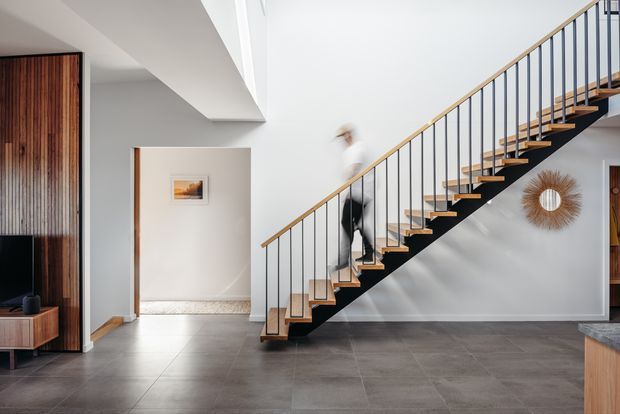 A bright contemporary interior focused on a floating staircase with black steel stringers, slim vertical balusters, and warm timber treads and handrail. The staircase rises through an open high-ceilinged space above a large-format grey tiled floor. White walls, timber built-in joinery, a small framed artwork, and a circular textured wall mirror complete the scene.