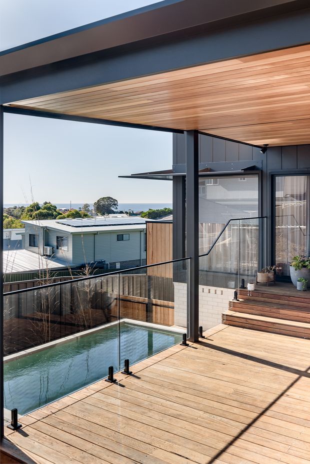 A sunlit elevated timber deck and balcony overlooking a small swimming pool, neighbouring homes, greenery, and a distant coastal horizon. The deck has clear glass balustrades with black metal posts and handrails, dark framed glazing, timber-clad soffits, and steps leading to a covered outdoor entertaining area with potted plants.