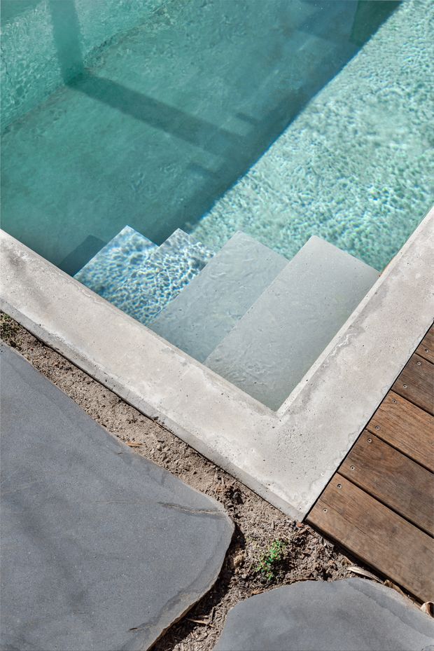 Close-up view of a modern swimming pool corner with broad submerged entry steps, pale concrete coping, clear blue-green water, and surrounding timber decking and dark stone paving. The image focuses on pool construction details and the transition between pool, deck, and landscape surfaces.