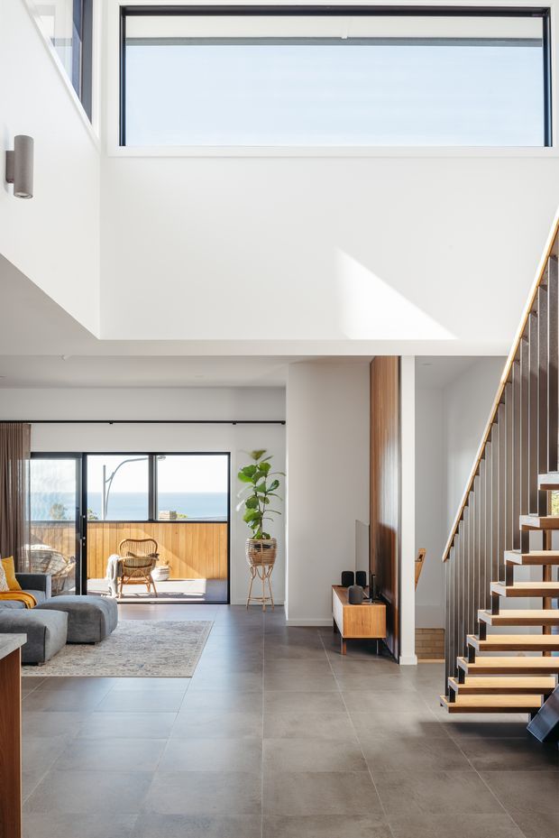 Bright contemporary open-plan interior viewed from the entry or circulation area, featuring a double-height void, floating timber staircase with dark metal balustrade, large grey floor tiles, white walls, timber joinery, and expansive glazing opening toward a coastal view. A living area with sofa, rug, armchair, and sliding doors is visible in the background.