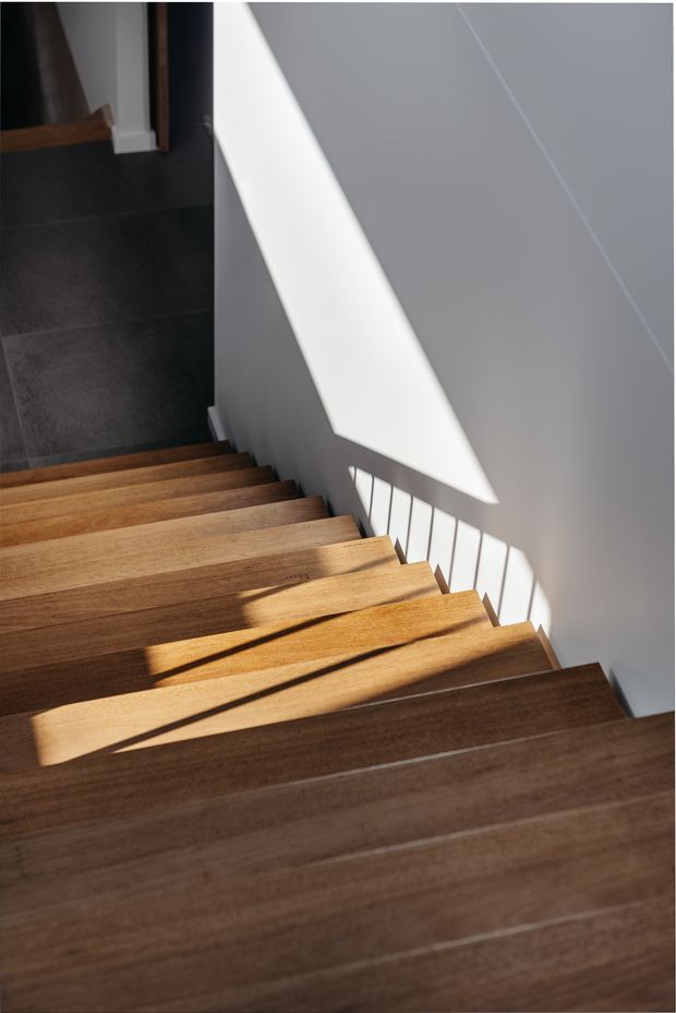 A contemporary interior stairway viewed from above, with broad warm timber treads descending toward a dark tiled landing. White and grey walls are illuminated by strong natural sunlight, casting geometric shadows from the stair railing across the steps and wall.