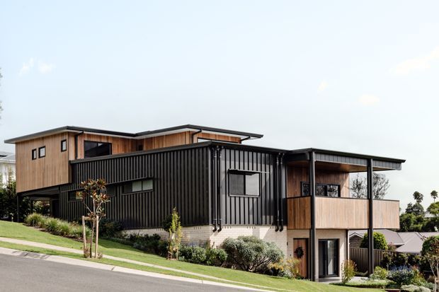 Exterior view of a contemporary multi-level residential house on a sloping suburban site. The building features dark vertical corrugated cladding, warm timber wall sections, pale masonry at the lower level, broad flat roof planes, black-framed windows, and a covered upper balcony or terrace.
