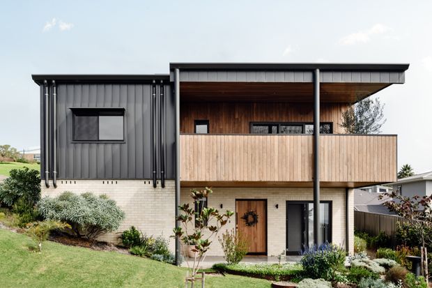 Modern two-storey residential house with a strong rectilinear composition, dark vertically ribbed metal cladding on the upper left volume, pale brickwork at ground level, and warm timber-lined soffits and balcony screening. A broad cantilevered upper terrace and flat roof create a sheltered outdoor living area, with landscaped garden beds and a sloping lawn in front.