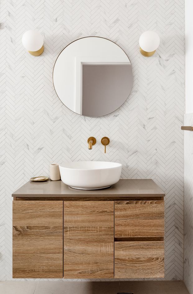 A refined contemporary bathroom vanity viewed straight on. A white vessel basin sits on a taupe stone-look benchtop above a warm natural timber floating vanity with drawers and doors. The wall is clad in white marble-look herringbone tile, with a large round mirror, brass wall-mounted tapware, and matching globe wall sconces.