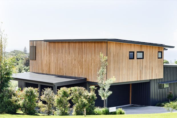 Exterior view of a contemporary multi-level house with a long, low-pitched roof and projecting eaves. The upper volume is clad in warm vertical timber boards with small dark-framed windows, while the lower level features dark ribbed cladding and a covered entry or carport-like structure. Landscaped planting and a sloping residential site surround the building.