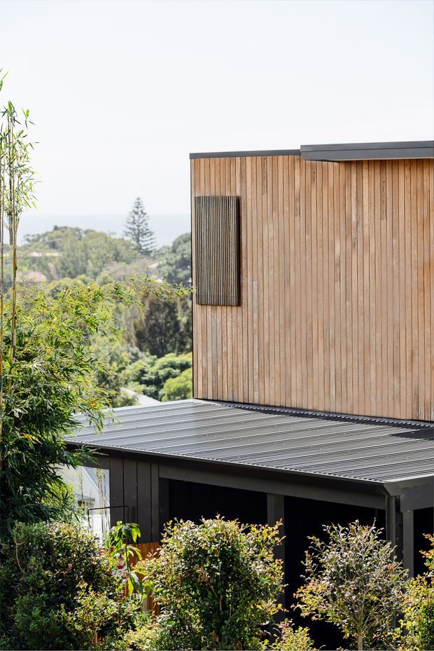 Exterior view of a modern multi-level building set within dense landscaped greenery. The structure features vertical timber cladding on the upper volume, dark metal roofing and trims, and dark lower-level cladding, with a distant wooded landscape visible beyond.