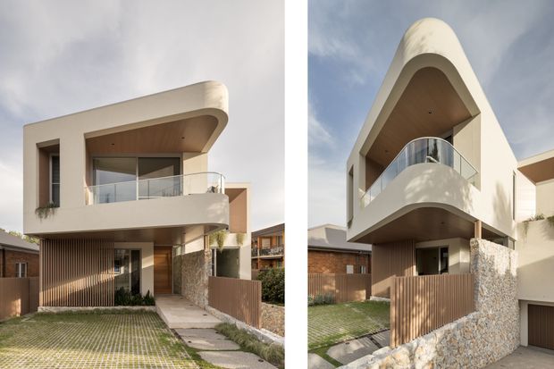 Two exterior views of a contemporary multi-level residence with sculptural rounded balconies and curved roof forms. The facade combines smooth light render, warm timber soffits and vertical timber screening, clear glass balustrades, and a textured natural-stone boundary wall. Small landscaped front gardens and pedestrian approaches are visible.