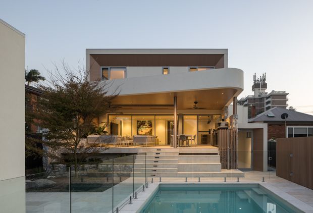 Contemporary multi-storey urban residence viewed from the rear, with a broad curved balcony and deep covered terrace above glazed living areas. Wide steps lead down to a rectangular swimming pool enclosed by frameless glass balustrading. The facade combines smooth light render, dark timber-toned soffit detailing, extensive glazing, and integrated warm lighting. Dense neighbouring buildings and rooftop equipment indicate a suburban or urban setting.