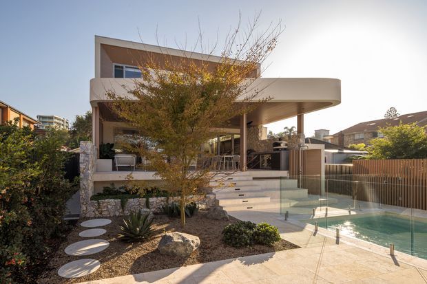 Exterior view of a contemporary multi-level residence with a broad curved cantilevered upper terrace, warm rendered walls, extensive glazing, stone-clad retaining elements, and an outdoor entertaining area overlooking a swimming pool. The landscaped front garden includes sculptural planting and stepping-stone pavers, while a timber privacy fence encloses the pool zone.