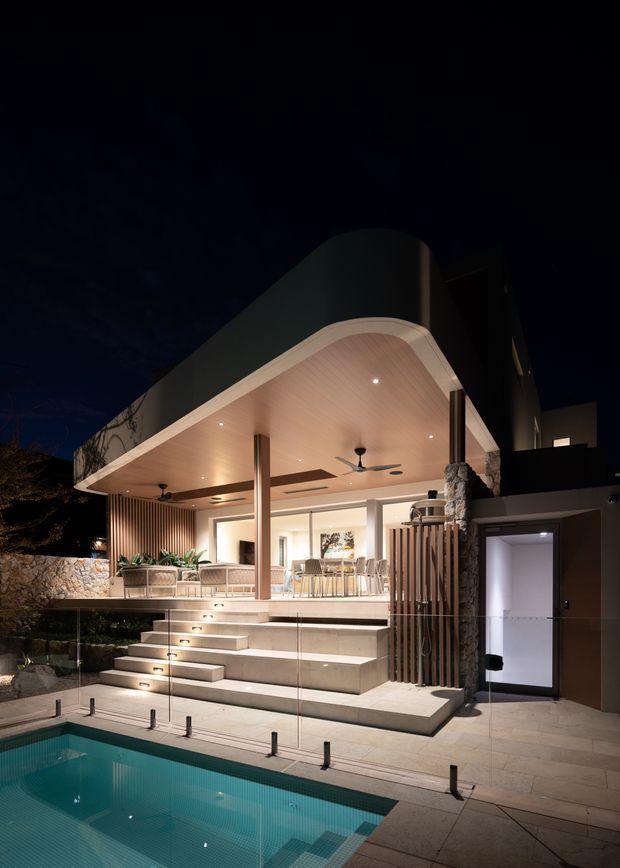 Night-time exterior view of a contemporary multi-level residence with a broad curved-roof outdoor entertaining pavilion overlooking a swimming pool. The covered terrace includes an outdoor lounge, dining area, ceiling fans, recessed lighting, timber-look soffit lining, stone columns and vertical timber screening. Wide tiled steps lead down to the pool, with glass pool fencing and landscaped stone retaining walls.