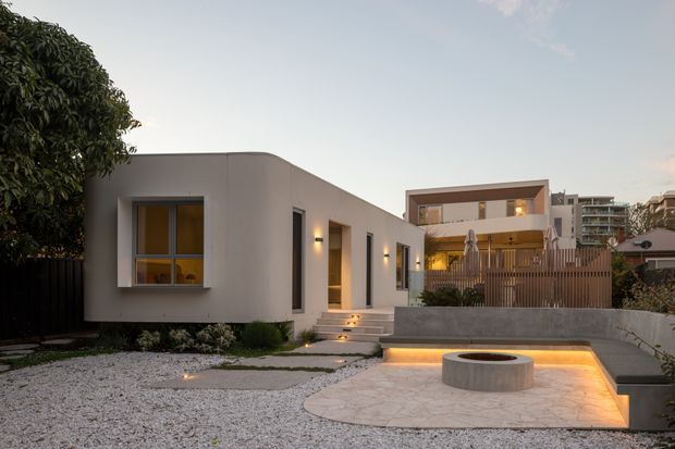 Twilight exterior view of a minimalist, single-storey flat-roof residence with smooth light-coloured rendered walls, simple rectangular windows, warm wall lighting, and illuminated entry steps. A pale gravel landscape and paved courtyard surround a circular built-in fire pit, with a raised timber privacy fence and neighbouring multi-storey buildings in the background.