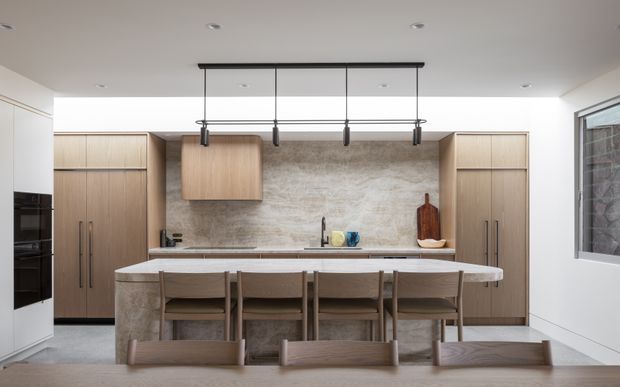 Contemporary kitchen viewed straight on, featuring a large stone-clad island with integrated seating, full-height light timber cabinetry, a textured stone splashback, concealed appliances, and a linear black pendant light. The palette is warm and neutral with clean, streamlined detailing.