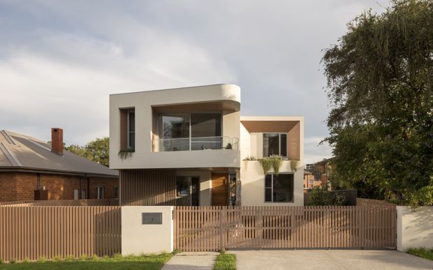 Street-facing view of a contemporary multi-level residence with a warm white rendered facade, curved upper-storey corner, recessed balcony, large glazed openings, vertical timber screening, and a slatted timber front fence. The home sits in an established suburban setting beside a brick house, with mature trees and a concrete footpath visible.