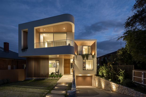 Twilight exterior view of a contemporary multi-level residence with a rounded projecting upper balcony, extensive glazing, warm recessed lighting, timber-lined soffits, vertical timber screening, and a garage accessed from a paved driveway. The home is set in a suburban landscaped setting with stone retaining walls and fencing.