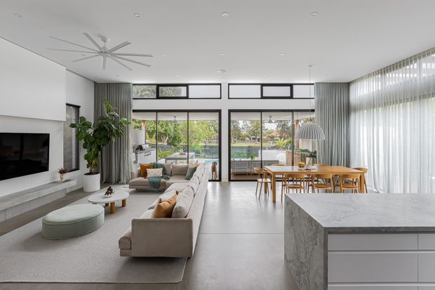 Open-plan contemporary living, dining and kitchen interior with polished concrete flooring, expansive floor-to-ceiling glazing, sheer curtains, a large stone kitchen island, a wall-mounted television, and light neutral furnishings. The bright room opens to a landscaped outdoor area and pool beyond the glazing.