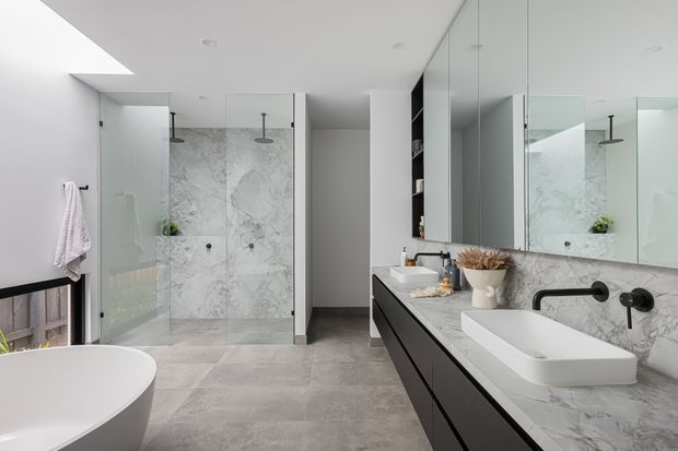 Contemporary bathroom with a freestanding white bathtub, large walk-in glass shower, double vanity with dark cabinetry, white stone benchtop, mirrored wall cabinets, and marble-effect wall surfaces. A skylight and bright neutral palette create an airy, refined atmosphere.