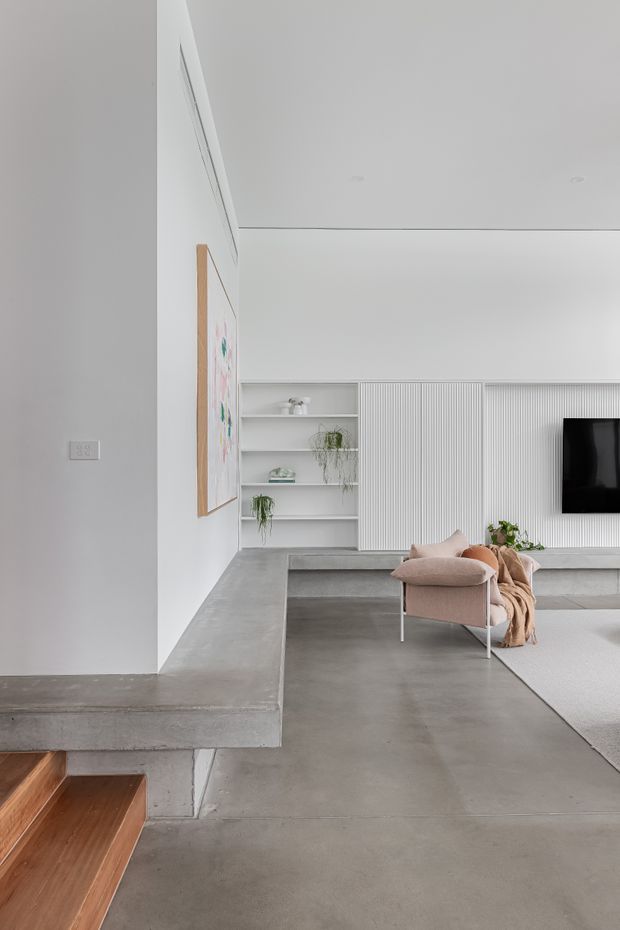 A minimalist contemporary living room with a polished concrete floor and integrated concrete hearth or platform beside timber stair treads. White walls and ceiling frame built-in open shelving, a vertically fluted media wall with mounted television, and a soft blush armchair with a draped throw. The room has clean lines, restrained neutral tones, and recessed ceiling lighting.