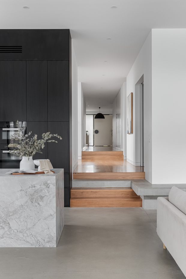 A refined open-plan residential interior viewed from a living area toward a hallway and entry. The scene features a dark full-height kitchen joinery wall, a veined stone island, polished concrete flooring, warm timber steps, white walls, recessed ceiling lighting, and a minimalist black pendant near the far entry. A light upholstered sofa is partially visible in the foreground.
