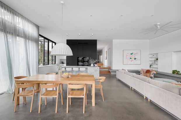 A bright, open-plan contemporary interior combining a dining area, kitchen and living room. The space features polished grey concrete flooring, white walls and cabinetry, a dark kitchen island backdrop, extensive sheer-curtained glazing, a timber dining table with matching chairs, a large white pendant light, and a pale sectional sofa. A stairway and built-in shelving are visible in the background.