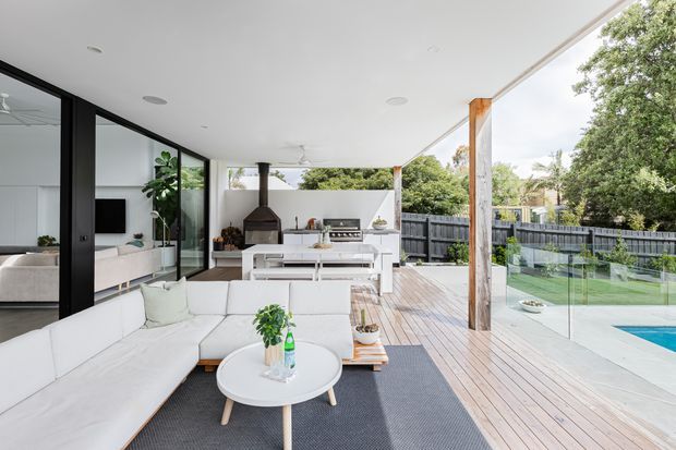 A bright, spacious covered outdoor entertaining area connected to a contemporary home, with a white sectional lounge, outdoor dining table, built-in barbecue, and freestanding fireplace. Timber decking extends around a glass pool balustrade, with the living room visible through large black-framed glazed doors.