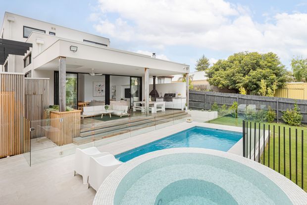 Contemporary multi-level home with a white rendered facade, broad flat roof overhangs and extensive glazing, viewed from a landscaped backyard. A paved entertaining terrace steps down to a bright blue swimming pool and circular spa, with a covered outdoor living area furnished with seating and an outdoor kitchen or barbecue zone. Timber screening, fencing and a manicured lawn frame the private pool area.