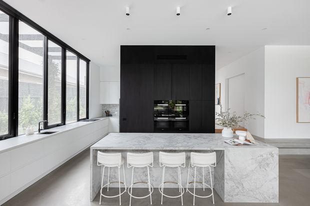 Bright contemporary kitchen with a large marble-effect island and four white bar stools, white perimeter cabinetry, black-framed floor-to-ceiling glazing, and a dramatic dark timber kitchen wall incorporating built-in appliances. The space has a clean, refined monochromatic palette and abundant natural light.