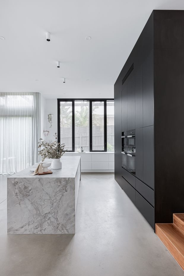 A bright, minimalist contemporary kitchen with a large veined stone island, full-height matte-black cabinetry and integrated appliances. Black-framed windows open the room to natural light, while sheer curtains, white walls, and a smooth grey concrete-like floor create a clean, refined interior. Timber stairs are visible at the right edge.