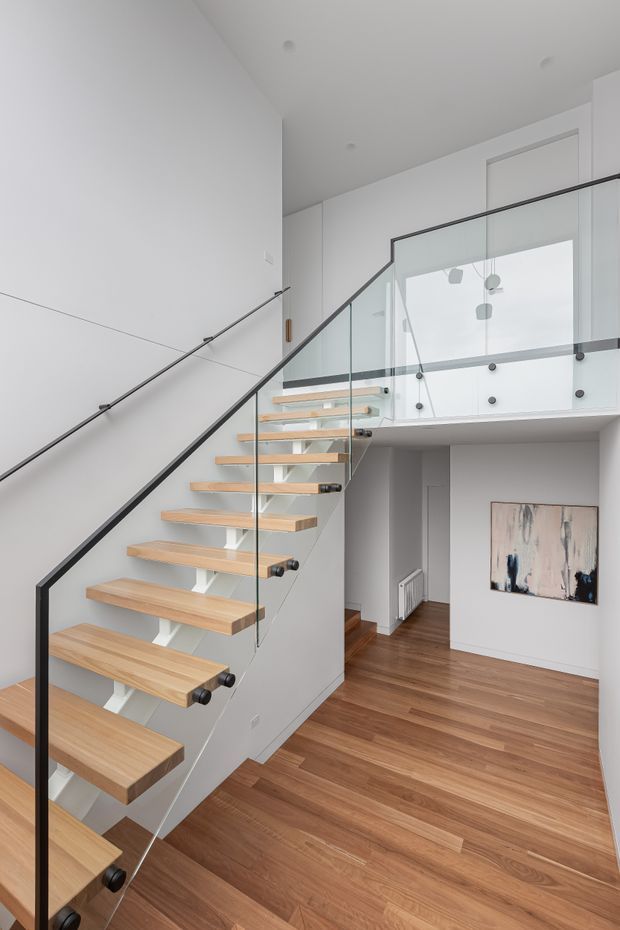 Bright minimalist interior view of a floating timber staircase with white supporting structure, black metal handrail, and clear glass balustrade. The stair rises to a mezzanine or upper landing overlooking a double-height hallway, with warm timber flooring, white walls and cabinetry, recessed ceiling lights, and a small abstract artwork.