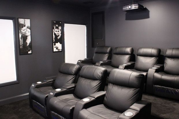 A dedicated home cinema room with several rows of black leather reclining theater seats, dark painted walls, framed film-themed artwork, carpeted flooring, and a ceiling-mounted projector.