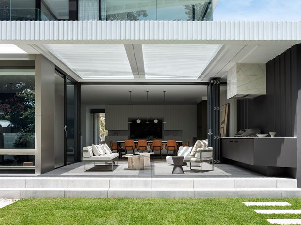 Contemporary open-plan indoor-outdoor living area viewed from the lawn, with large glazed openings, a covered terrace, outdoor lounge seating, dining table, and a dark built-in kitchen/bar at the rear. White ribbed facade detailing, pale tiled paving, and strong black-and-white contrast define the design.