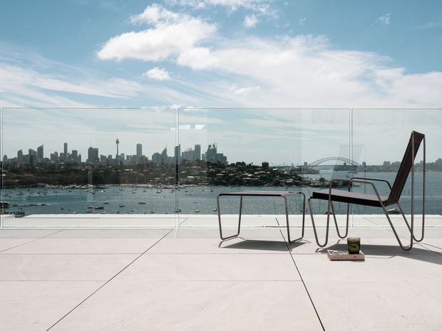 A minimalist rooftop terrace or balcony with pale tiled paving and transparent glass balustrades, overlooking a harbour with boats and a prominent city skyline and bridge. Sparse modern outdoor furniture occupies the foreground, creating an open, urban waterfront setting under a bright partly cloudy sky.