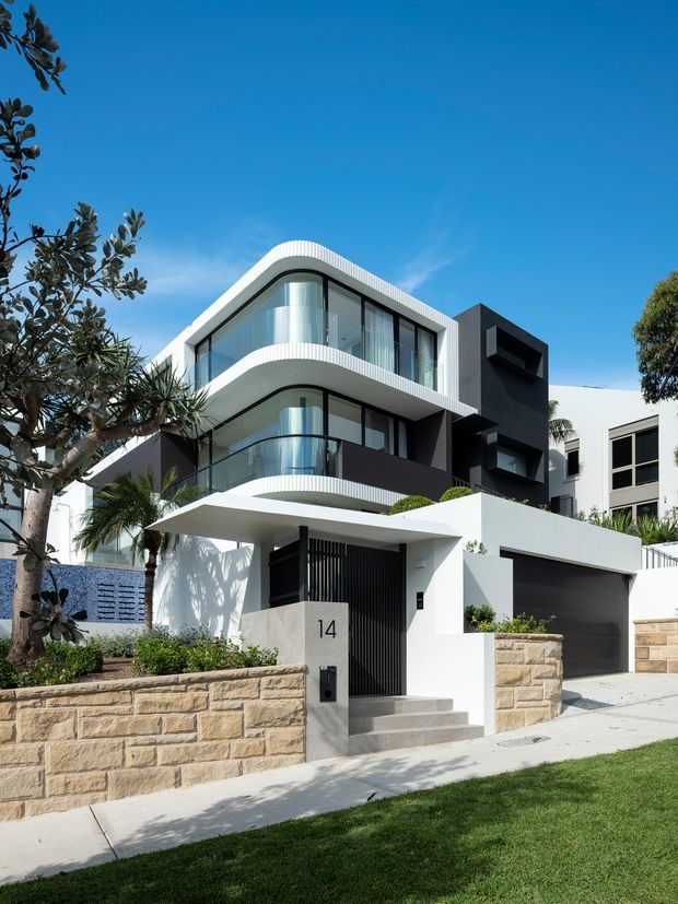 Street-level exterior photograph of a contemporary multi-storey urban residence with rounded projecting balconies, extensive curved glazing, white rendered forms, dark charcoal cladding, a flat roofline, integrated garage, gated entry, and sandstone boundary walls. Landscaped planting and lawn frame the modern facade.