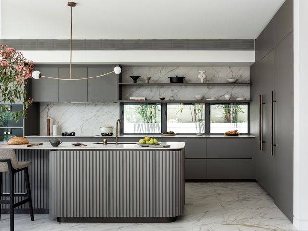 A refined contemporary kitchen featuring a large fluted-front island with seating, dark grey flat-panel cabinetry, a pale veined stone benchtop and matching floor, open marble-look shelving, and integrated appliances. A linear sculptural pendant hangs over the island, while clerestory glazing and windows provide natural light.